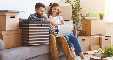 Happy couple sitting with a laptop amongst moving boxes in their new home. 